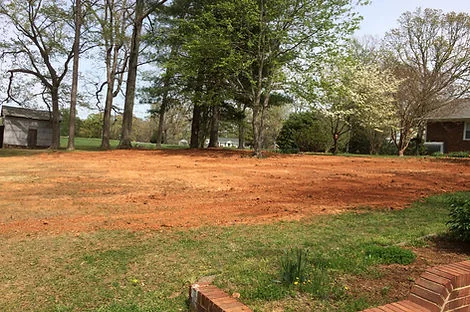 Cleared red clay lot with mature trees and a house in the background