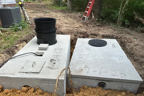 Two concrete septic tanks side by side with a black riser on one lid near an outdoor air conditioner Two concrete septic tanks side by side with a black riser on one lid near an outdoor air conditioner